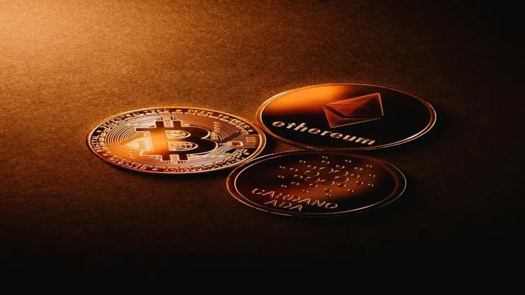 Imagem de três moedas físicas representando Bitcoin, Ethereum e Cardano (ADA) em um fundo escuro com iluminação dourada.